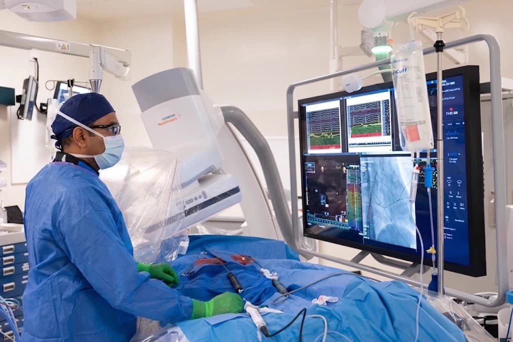An electrophysiology study in progress at Heart Health Center. A physician stands at the draped table with catheters entering the femoral access site, watching a monitor stack that displays intracardiac electrograms above a fluoroscopic image of the catheters in the heart.