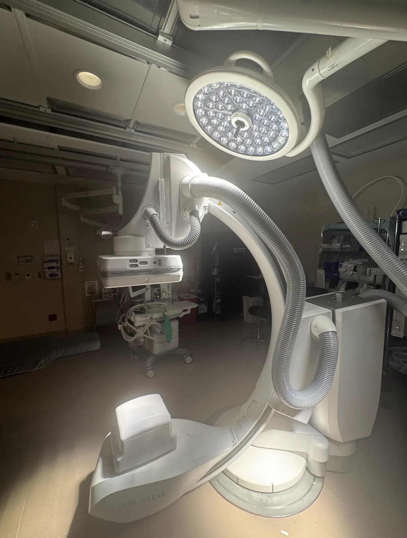 A close view of the fluoroscopic C-arm, its detector and X-ray source facing each other across the arc where the table sits, lit from above by a surgical light on an articulated boom.