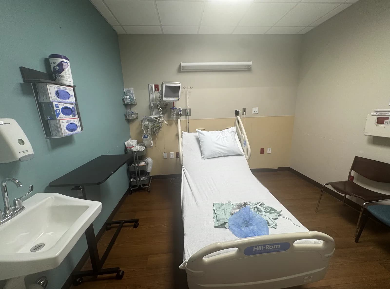 A pre-op bay with a bed, a wall-mounted monitor, a hand basin, a supply cart, and gloves and dressings stocked within reach at the bedside.