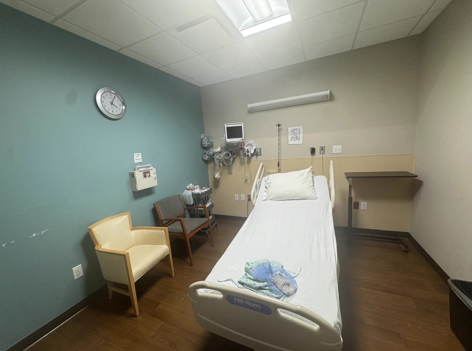 A private recovery bay: a hospital bed made up with a folded gown on it, a wall-mounted patient monitor and oxygen and suction outlets at the head of the bed, a clock on a teal accent wall, and a chair for a family member.